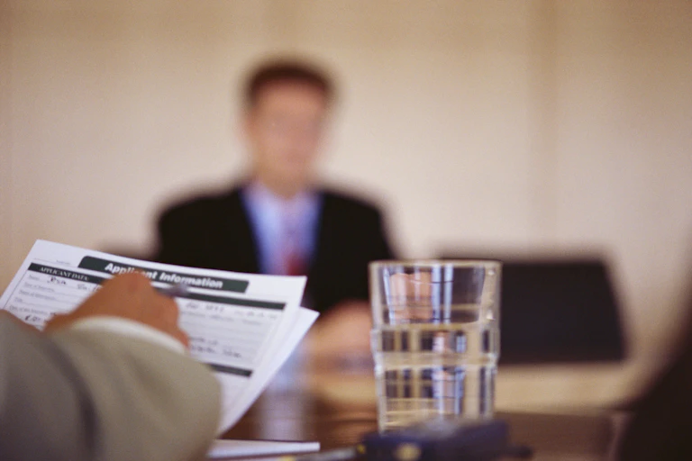 A man in a suit and tie, whose features are blurry, sits at a table across from someone holding a job application.