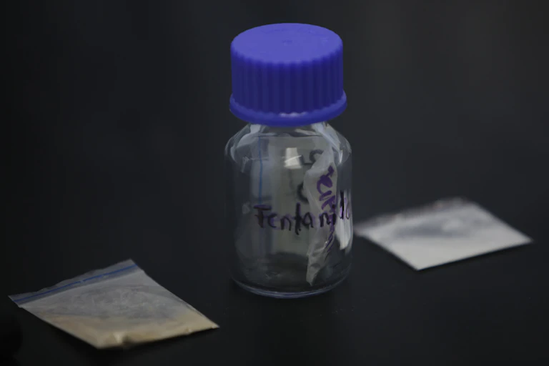 Glass vial with blue cap stands in between two cellophane baggies that contain powder