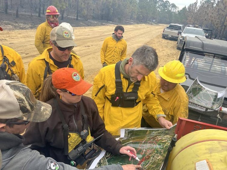 Eight people in yellow fire clothes look at a map. The air around them is smoky.