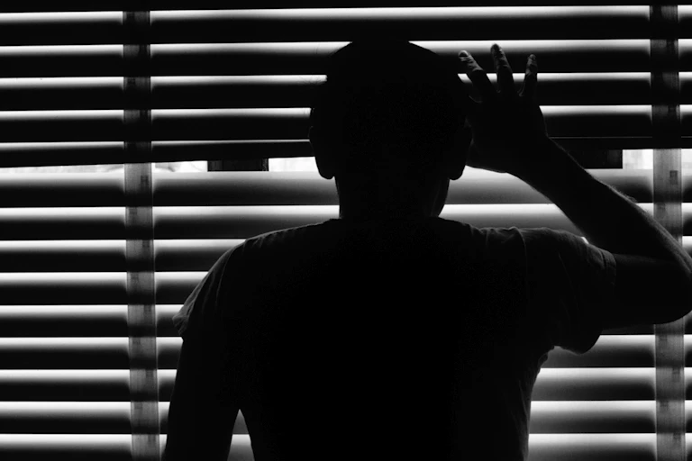 Silhouette of young man peering through window blinds.