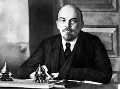 A black-and-white photo of a bald man with a moustache and goatee in a suit jacket.