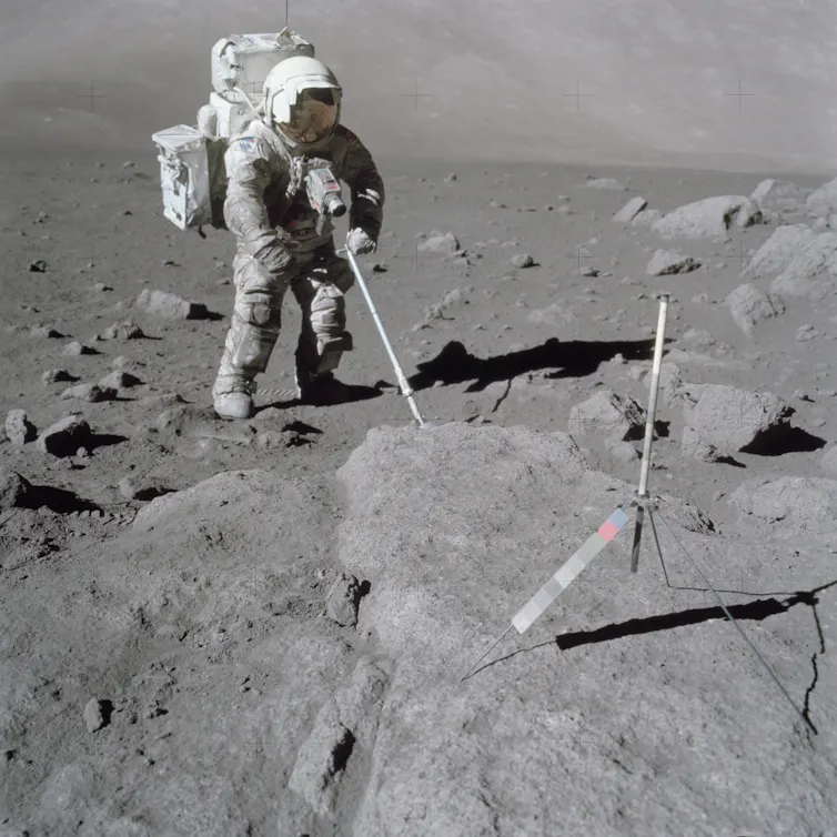 An astronaut standing on the lunar surface and using a scoop to retrieve samples.
