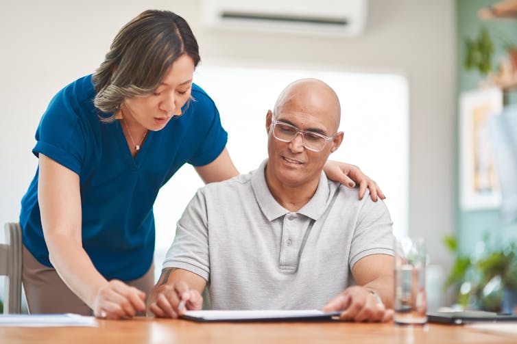 A woman in a blue shirt stands over a man who is sitting at a table filling out a document.