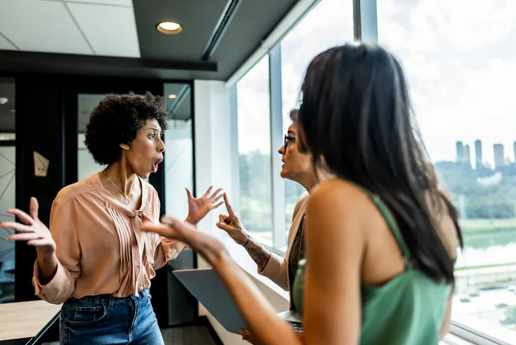 three women in an office gesticulate at each other heatedly