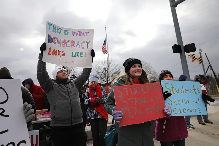 A group of people stand on the street holding handmade signs. One says 'This is what democracy looks like.'