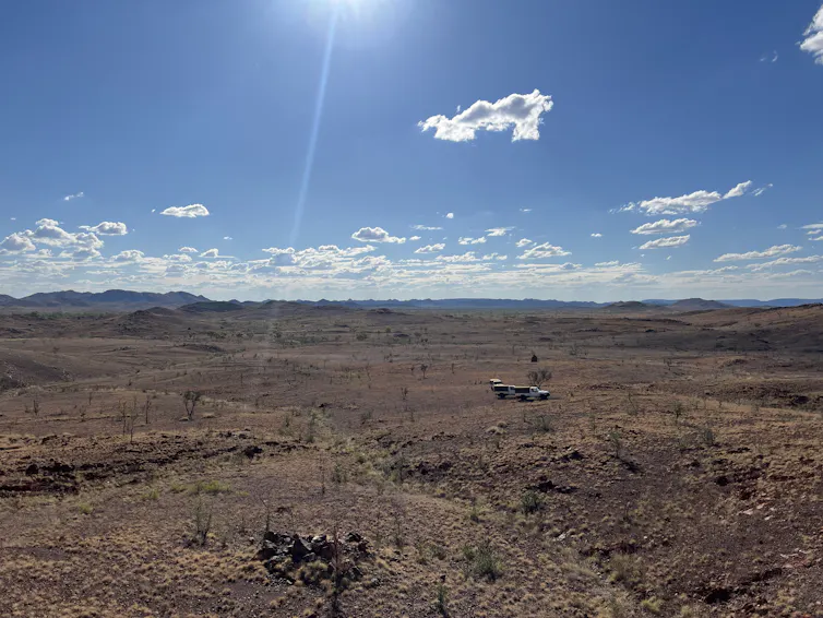 Uma visão ampla de uma paisagem avermelhada sob um céu azul com vários veículos 4x4 à distância.