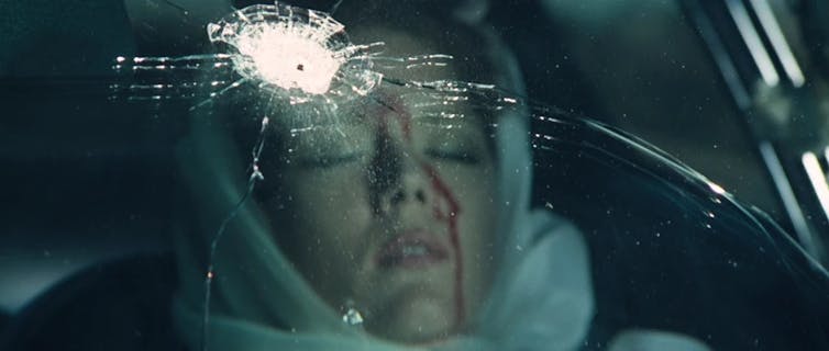 Screen grab of a young woman stiting in a car, with a bullet hole in the glass and blood trickling down her face.
