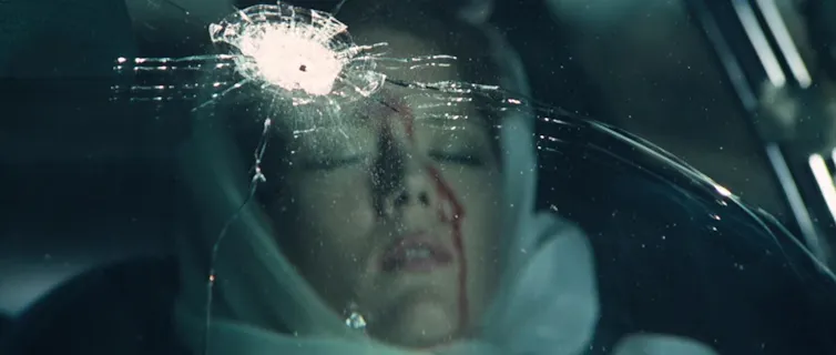 Screen grab of a young woman stiting in a car, with a bullet hole in the glass and blood trickling down her face.