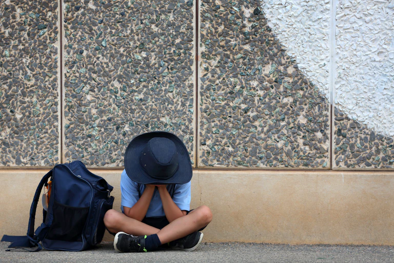 A young boy in a uniform sits with his head in his hands. A backpack rests next to him.
