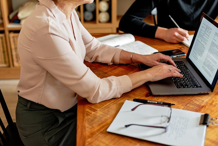 A woman sitting at a wooden desk typing on a computer, with a notepad by her side.
