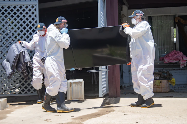 Three SES volunteers wearing personal protective equipment including masks remove flood-damaged furnitutre and a television from a property after a cyclone.