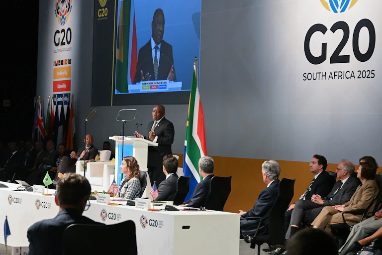 A man speaking at a podium, also seen on a big screen behind him. People seated at a long table are turning to look at him.