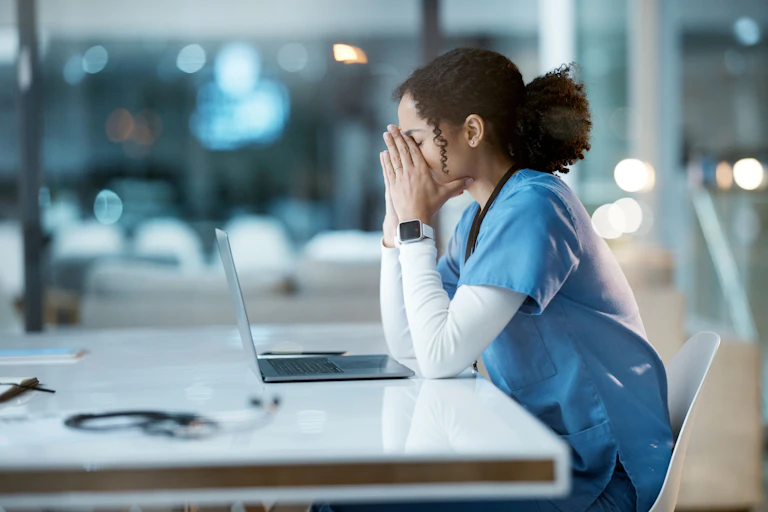 tired and stressed nurse sitting at a laptop with her head in her hands.
