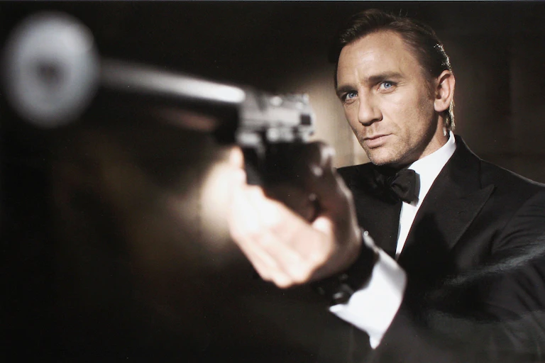 Man wearing tuxedo aims a pistol with a silencer affixed to it.