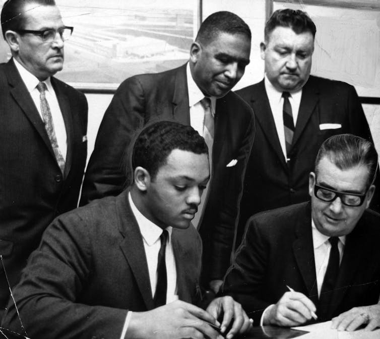 Jackson, in a suit, sits at a table next to a white man signing a document surrounded by other men in suits