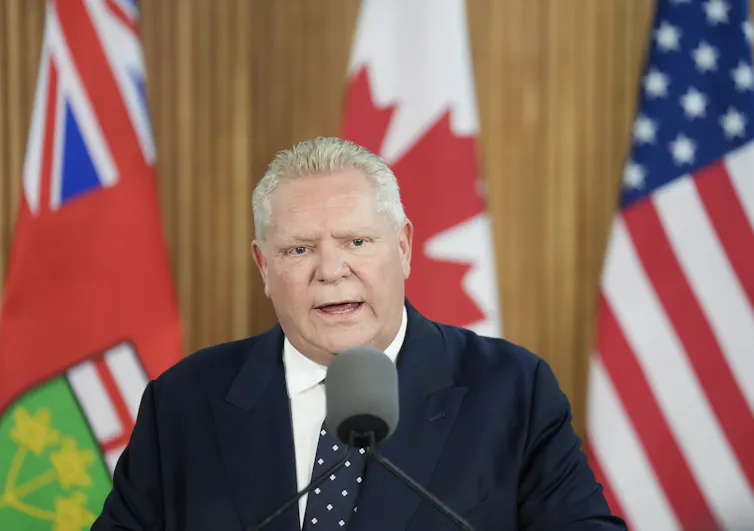 El hombre de Rotunde con retrolavado deslizante habla en un micrófono frente a Ontario, Canadá y banderas estadounidenses.