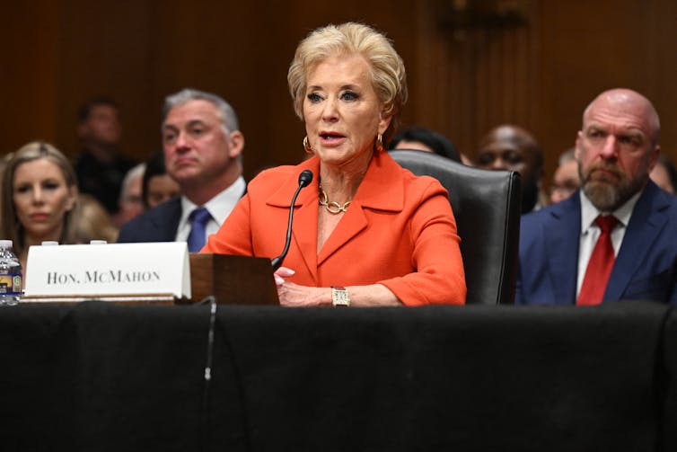A woman, dressed in a bright orange jacket, seated with several others, speaks from behind a desk.