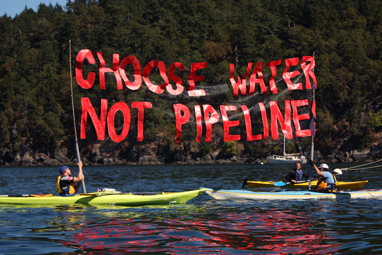 Kayakers on a lake hold up a banner reading 'Choose water, not pipelines'