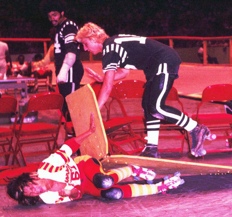 Female skater in red uniform lies on ground under wooden board as woman skater in black uniform tries to pass