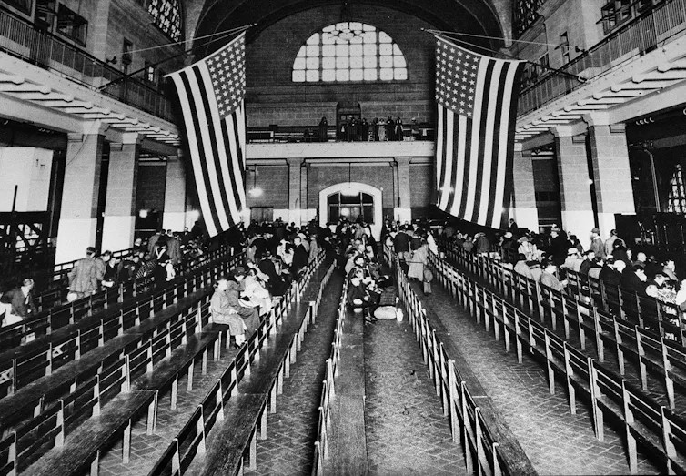 People sit in a large room with two American flags hanging in a black and white photo.
