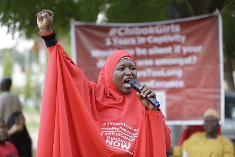 A woman wearing a red hijab speaks into a microphone and raises her arm while making a fist