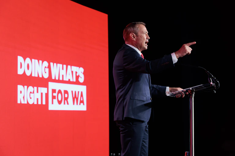 A man points and speaks in front of a sign that says doing what's right for WA