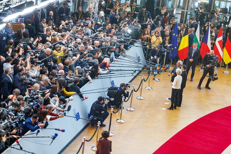 A birds-eye view of three people answering questions from a large press pack.