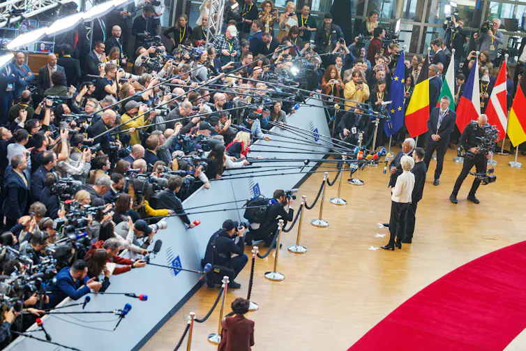 A birds-eye view of three people answering questions from a large press pack.