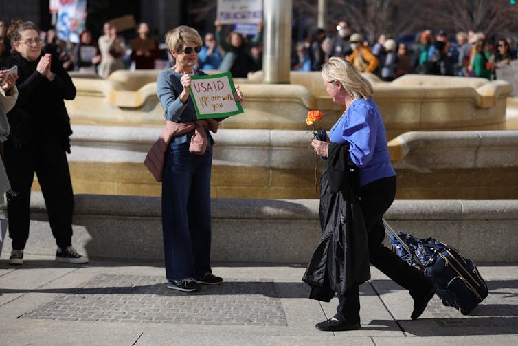 A blonde woman wearing a blue shirt and holding an orange flower walks past people and wheels a suitcase outside.