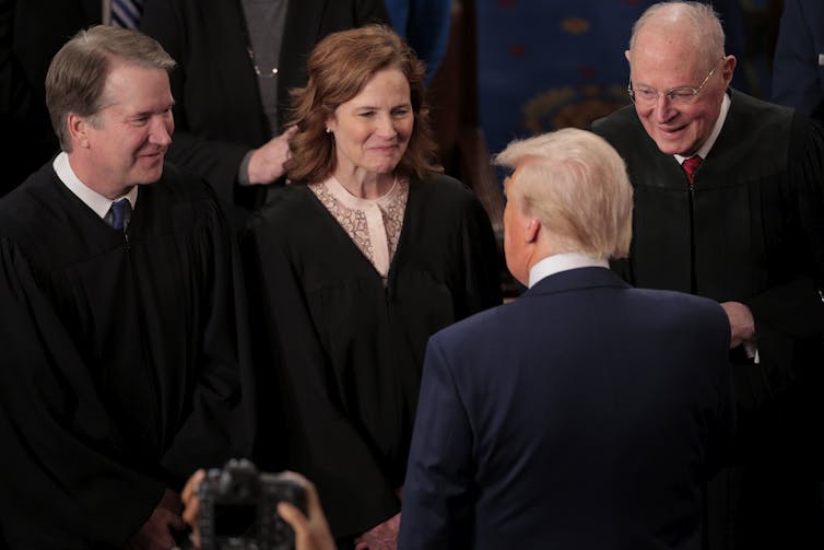 Two white men and a white woman all wearing black robes face a man with white hair and a dark suit and smile at him.