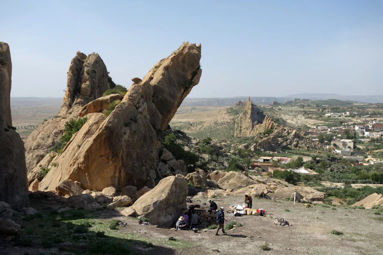 A rocky outcrop with people in white working at ground level beneath it; in the background white buildings from a village.