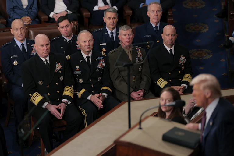 People in military uniforms look on as a man in a suit speaks at a lectern.