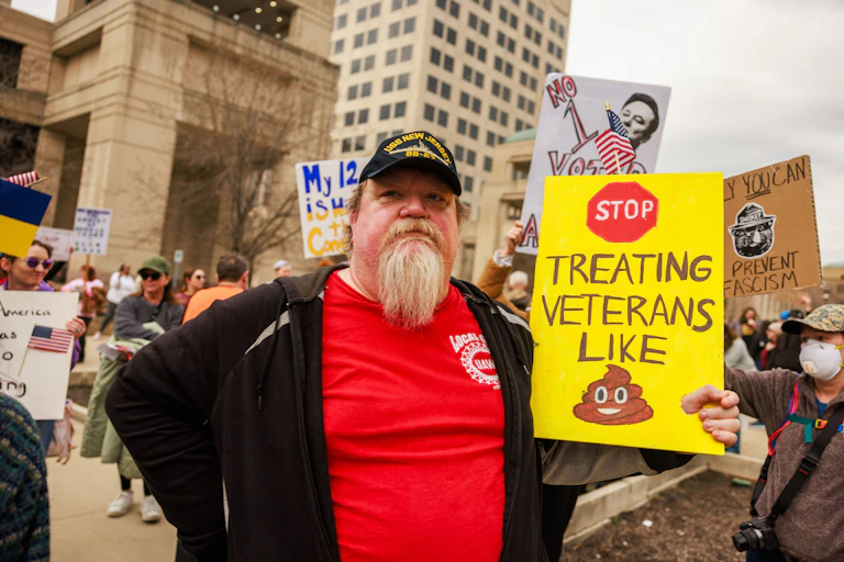 A protester holds a sign reading, "Stop treating veterans like shit" during a demonstration outside some large buildings.