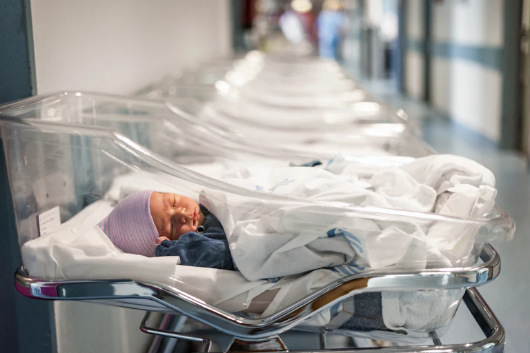 A newborn baby wearing a pink hat lays in a clear plastic bassinet in a hospital. A row of similar, empty bassinets are arranged alongside it.