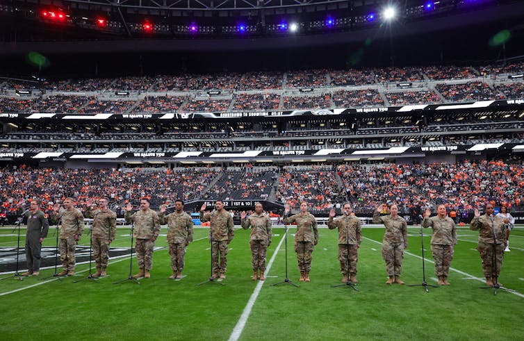 People in camouflage uniforms raise their right hands while standing on a field in a sports stadium.