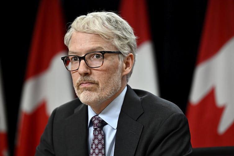 Un hombre blanco con gafas y cabello blanco con un traje oscuro sentado frente a las banderas canadienses en los pilares