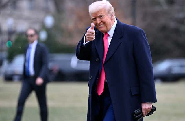 An old man with orange-tinged skin and whiteish hair wearing a dark suit and red tie gives a thumb's up to the camera