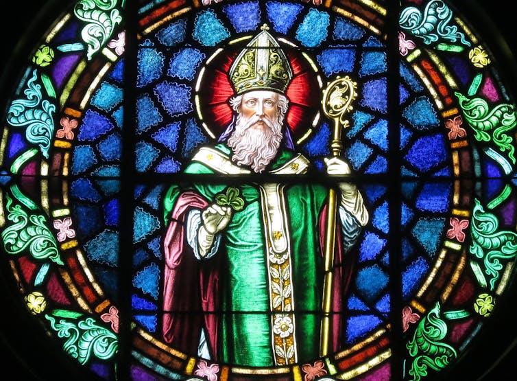 Stained glass window of bearded man wearing a green robe standing against a blue backdrop.