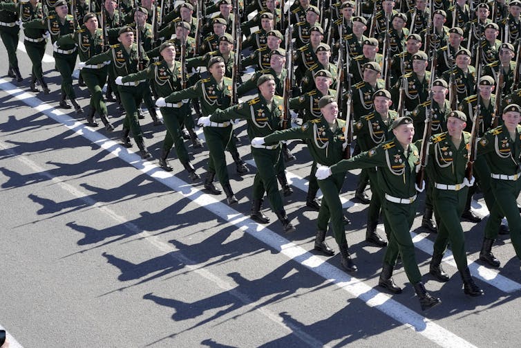 Men in green military uniforms march down a street in formation