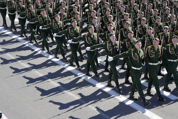 Men in green military uniforms march down a street in formation