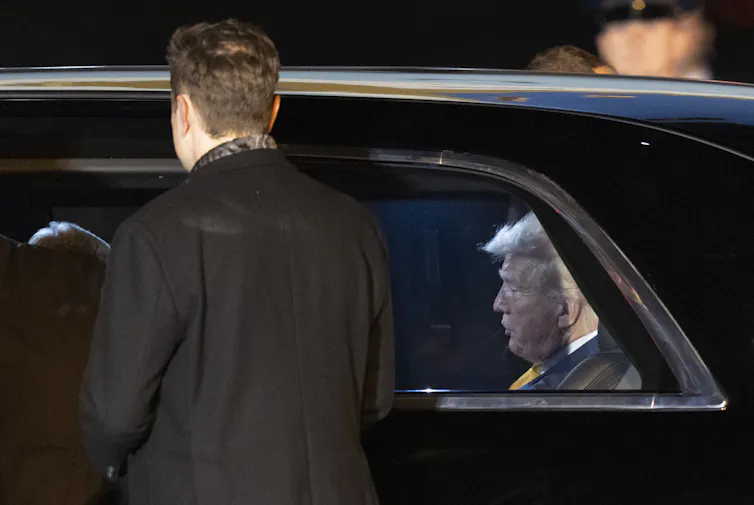 A man with white hair sit in the back of a limousine while a man with dark hair stands outside it.