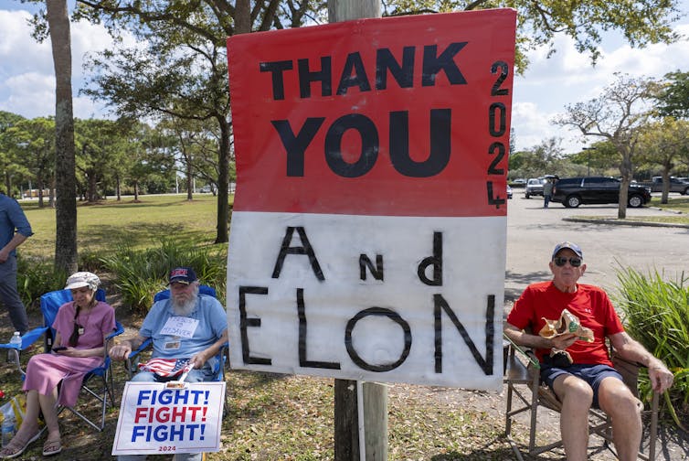 Elderly Trump supporters sit in chairs by the side of a road holding up a sign that reads Thank You and Elon.