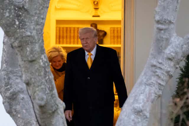 An older man with fluffy white-blond hair is photographed between two grey tree branches leaving an ornate office.