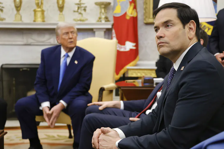 A younger man with dark hair sits looking away from another older man with fluffy white-blond hair and orange-tinged skin.