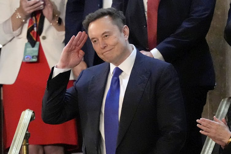 A dark-haired man in a dark suit salutes.