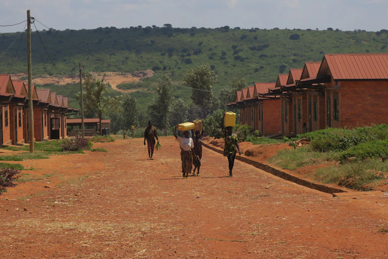 A dusty red road with two rows of houses on either side and people walking with 20 litre water buckets on their heads
