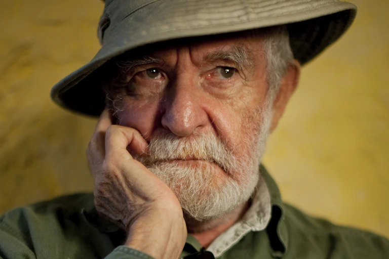 An elderly man with a grey beard and a khaki sun hat looks pensive, a hand to his jaw and his eyes looking into the distance.