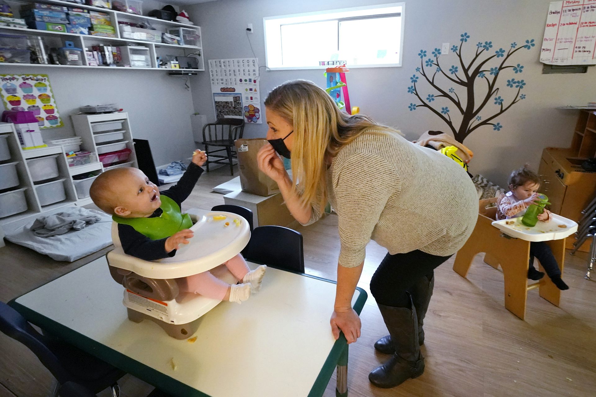 A baby sitting in a high chair reaches for their carer who is wearing a black COVID mask.  