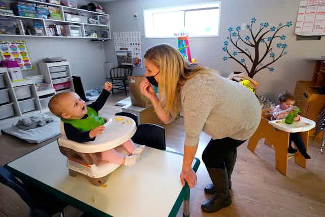 A baby sitting in a high chair reaches for their carer who is wearing a black COVID mask.