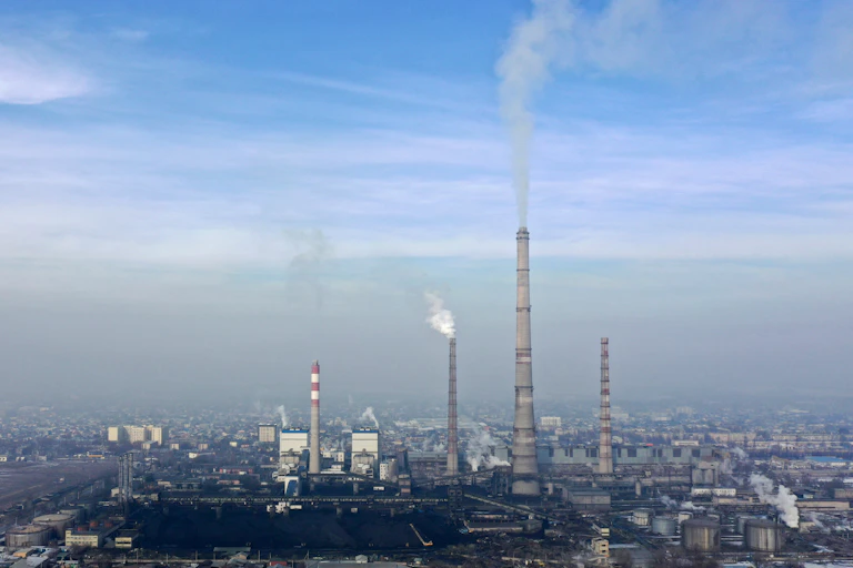 Tall facilities at a thermal power plant emit a gray substance over a city, with smog in the background.